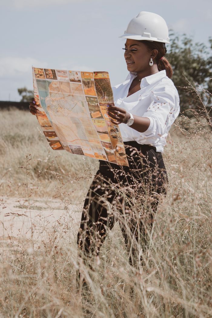 Services A female engineer in a safety helmet reads a map in a grass field on a sunny day.