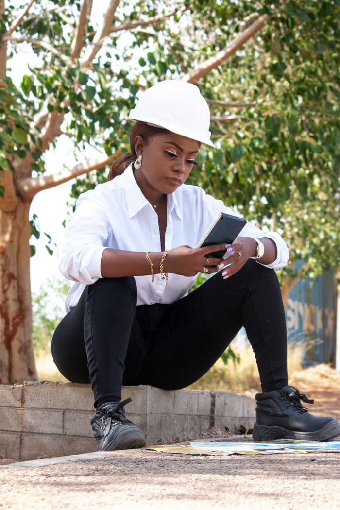Services African female engineer in hard hat using smartphone outdoors, emphasizing technology in construction.