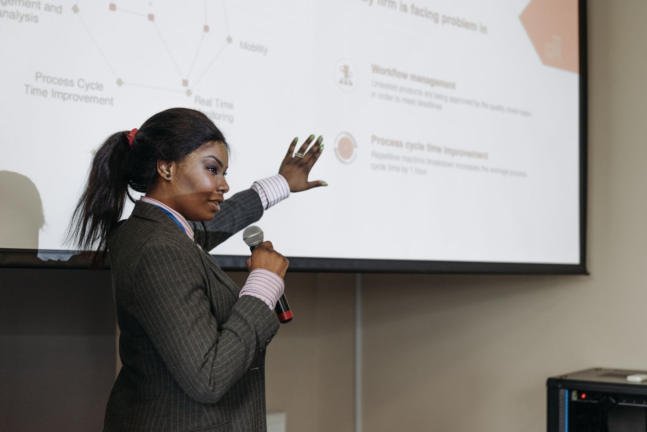 The Art of Drawing Readers In: Your attractive post title goes here Businesswoman presenting with microphone, discussing improvements with a slide projection in a conference room.